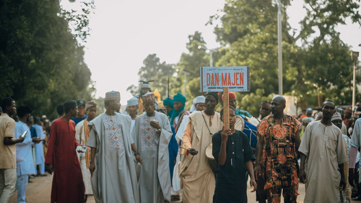 A vibrant cultural procession with a diverse group holding a 'Dan Majen' sign outdoors.